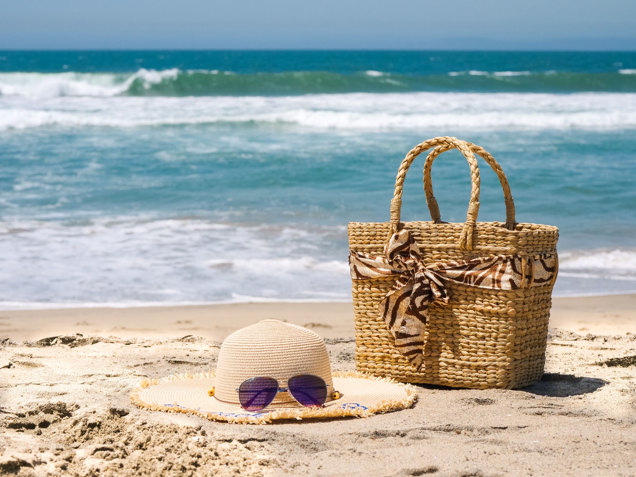 Straw hat, beach bag, and sunglasses on sandy beach with ocean waves.